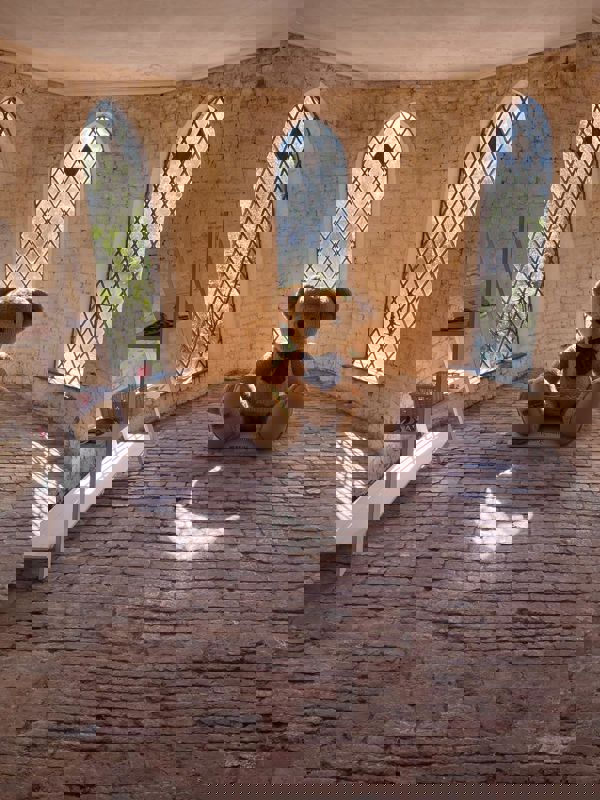 A life-sized teddy bear with  a dark blue bow around its neck sat on a cobbled floor with windows behind it.