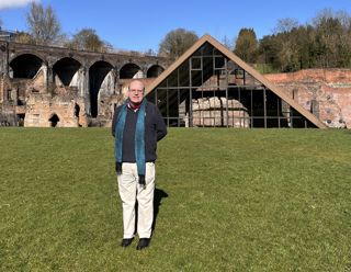 A smiling man in cream trousers and a navy jumper standing in front of a triangle-roofed building containing industrial ruins and with the ruins of industrial buildings in the background.
