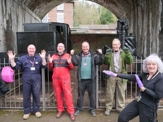 Four men in works overalls, smiling and with their arms raised in front of them as if they are celebrating something, standing outdoors in front of two locomotives under a brick arch and accompanied by a smiling woman pointing towards the men as if to draw your attention to them because they have done something good..