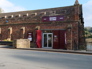 A brick building with crenellations and a museum entrance with signage in the foreground. In the background you can see a river.
